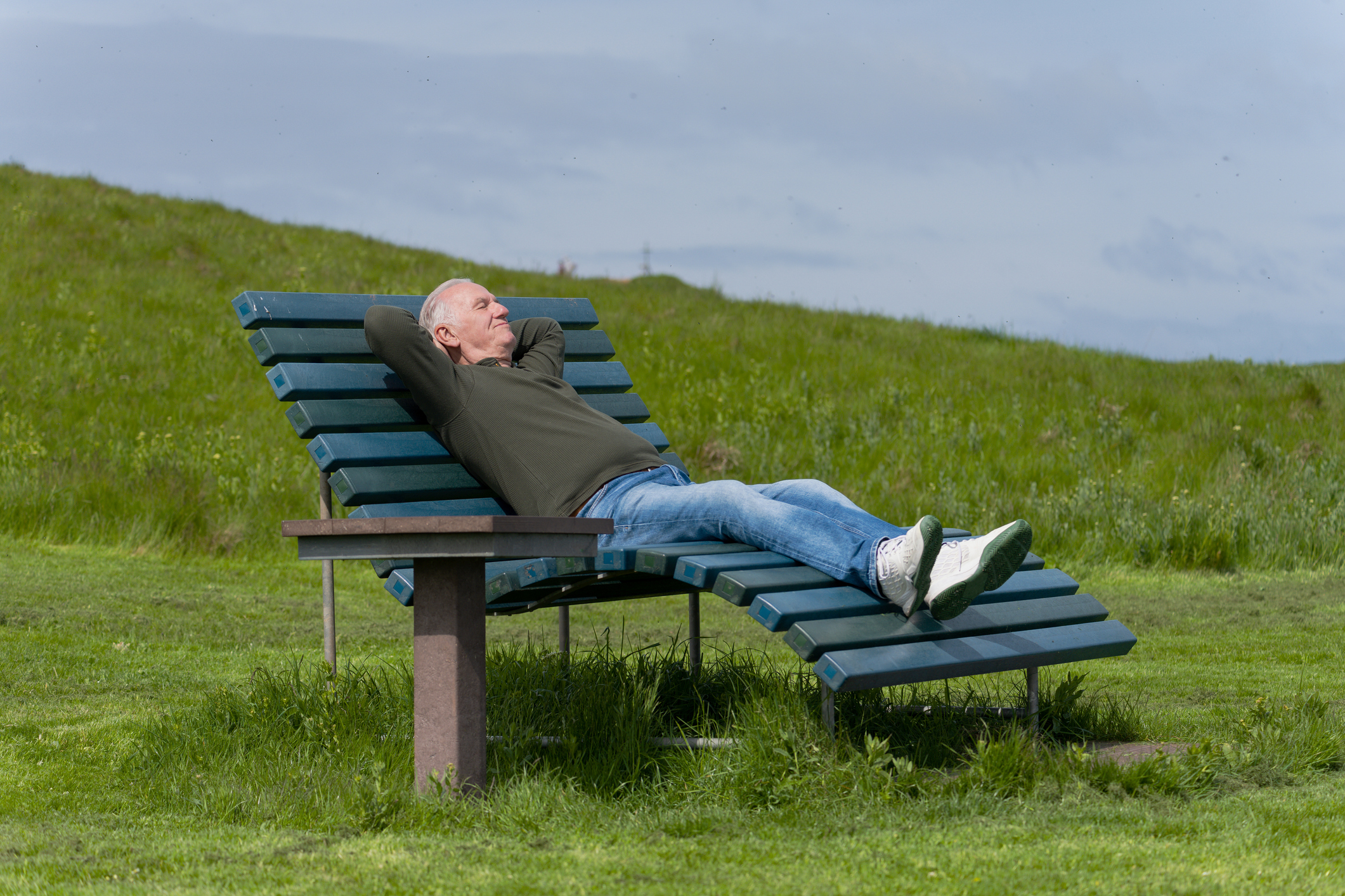 Älterer Mann, der auf einer Bank im Sonnenlicht auf einer Wiese liegt.